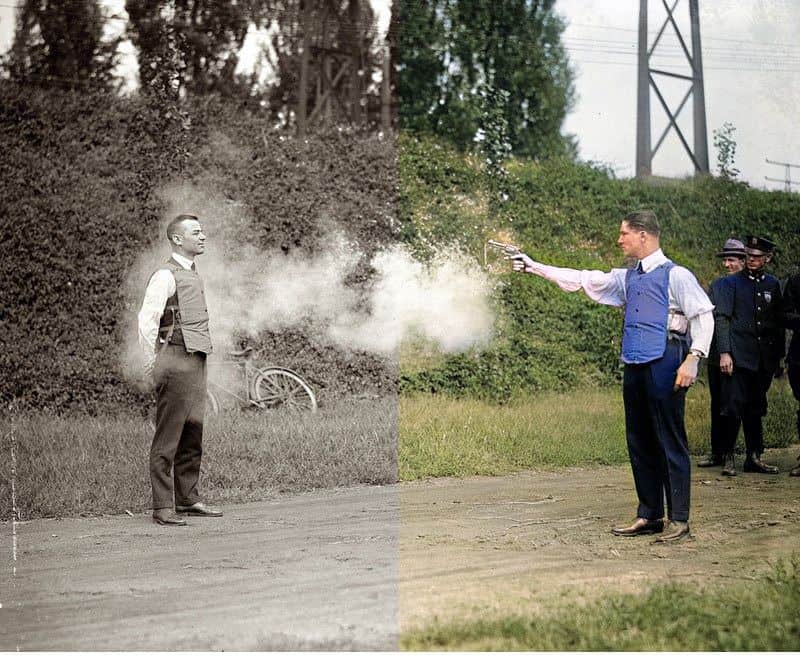 Man aiming a gun at another man in an outdoor setting.
